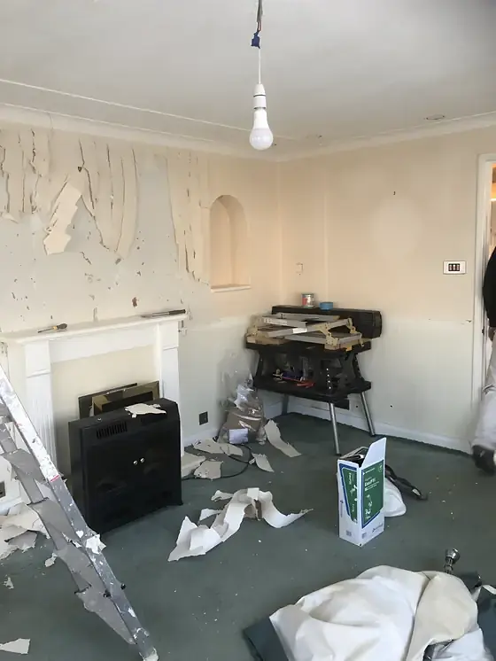 Room mid-strip with peeling wallpaper, scattered scraps, a stepladder and a white fireplace