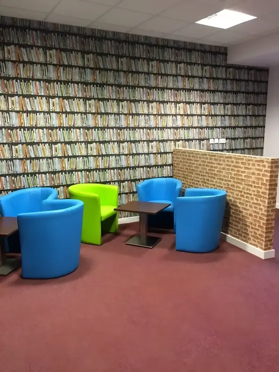 Student lounge in Colchester with bookshelf-print wallpaper, brick-effect partition and bright blue and green tub chairs