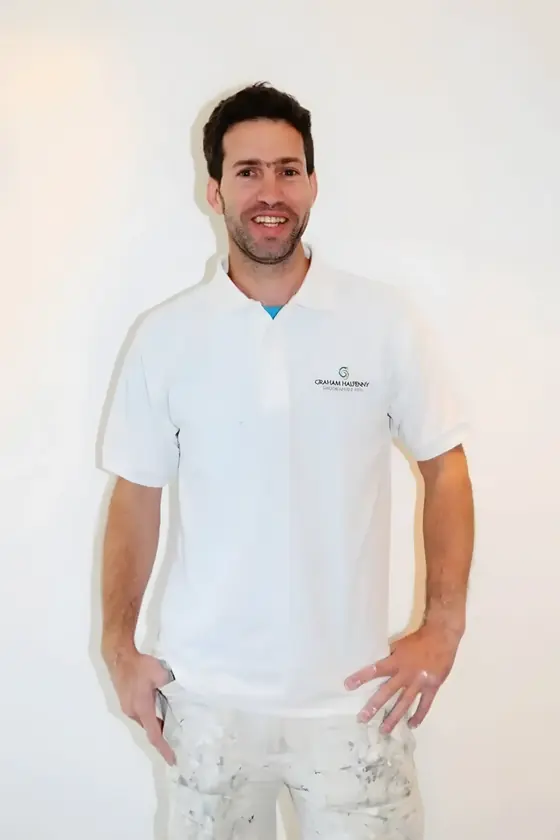 Smiling decorator in white polo shirt and paint-flecked trousers against a plain white wall