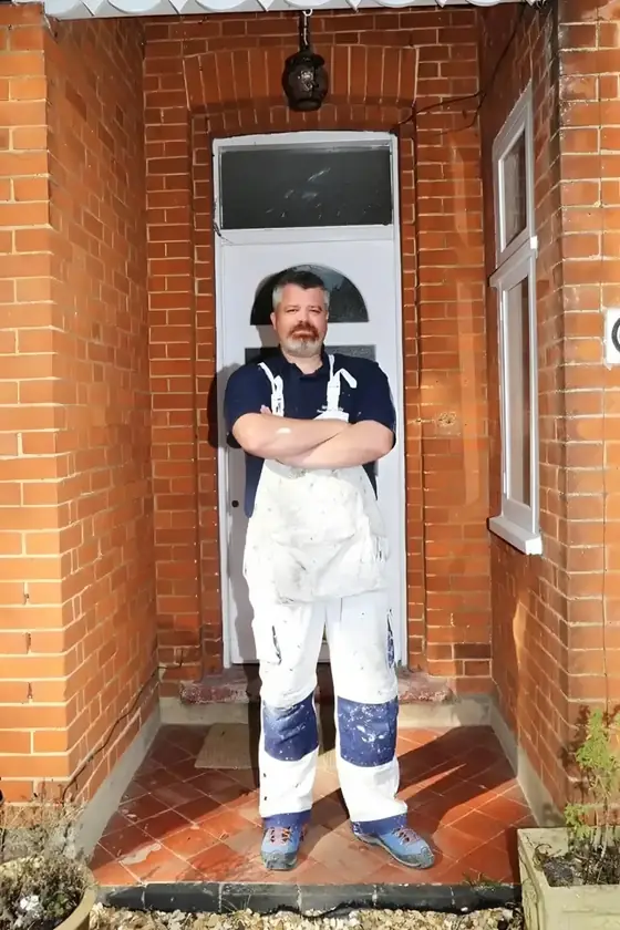 Painter with folded arms in paint-splattered white overalls in a red brick porch