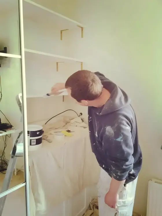 Decorator painting fitted shelving brackets and shelves in a bedroom, dust sheet over furniture