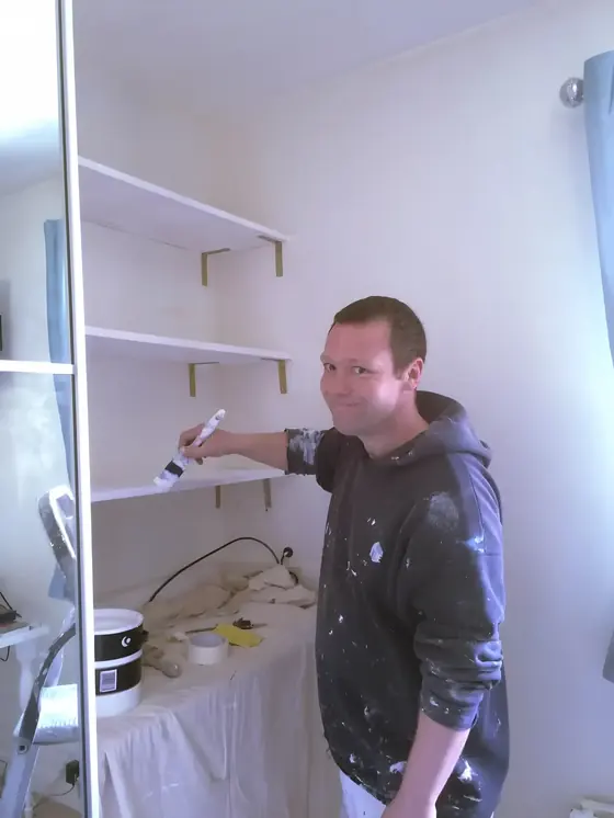 Smiling decorator painting built-in white shelves in a bedroom, paint tin below