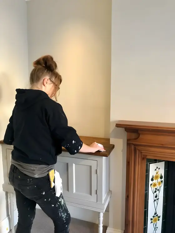 Painter in a black top rubbing down a grey painted cabinet beside a tiled fireplace
