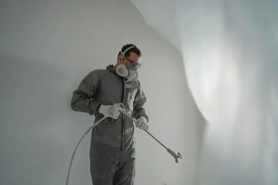 A person in a respirator and overalls holding an airless spray gun in a white room